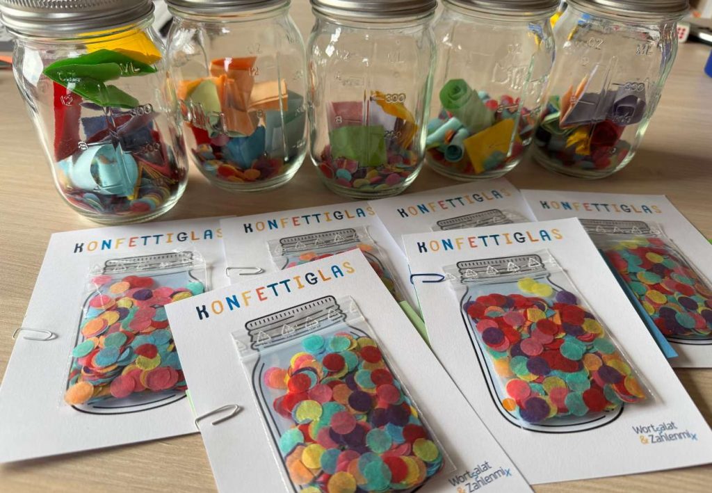 Gläser mit Konfetti uns kleinen bunten Zetteln im Hintergrund und Postkarten mit einem Konfettiglas und echtem Konfetti im vordergrund